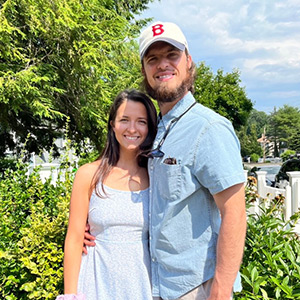 A husband wearing a light blue shirt with a Boston red sox cap puts his arm around his wife who is wearing a white dress on a sunny day.