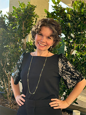 A patient in a black dress smiling with her hands on her hips standing front of several green plants